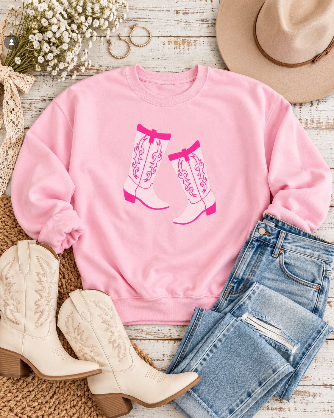 Pink cowboy boots sweatshirt styled with jeans, cream cowboy boots, a tan hat, hoop earrings, and white flowers
