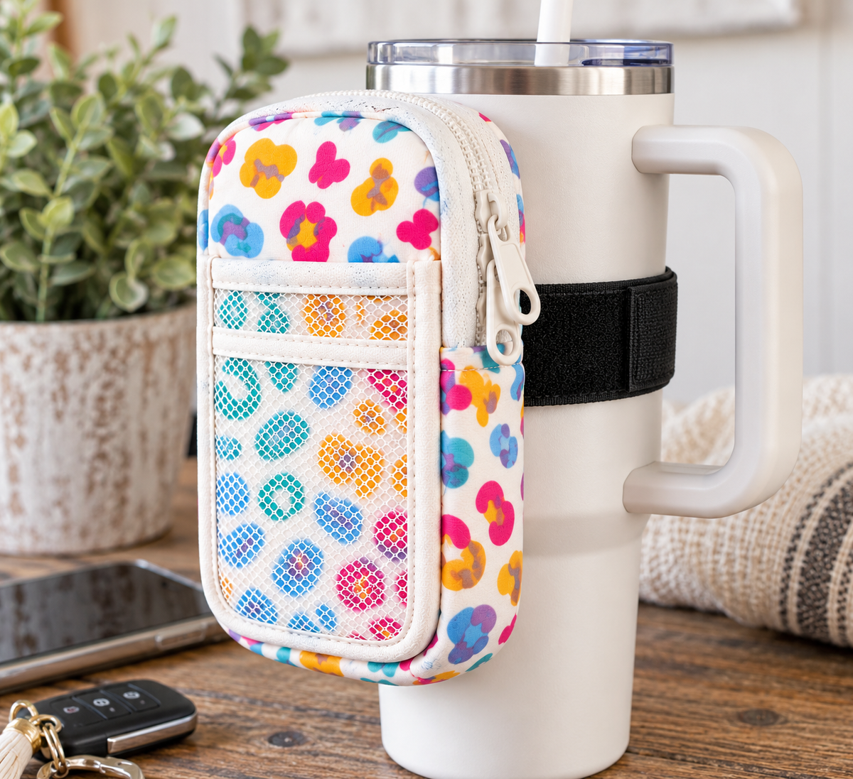 Multicolor Floral Tumbler Pouch attached to white Stanley tumbler handle with black elastic strap showing multicolor retro floral print in pink, teal, blue, orange, and purple, white side zipper, slip pocket, and mesh pocket on rustic wood desk beside car keys and potted greenery
