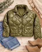Olive cropped quilted snap-front jacket styled with jeans, tan ankle boots, a cream fringed textile, and greenery on pale rustic wood
