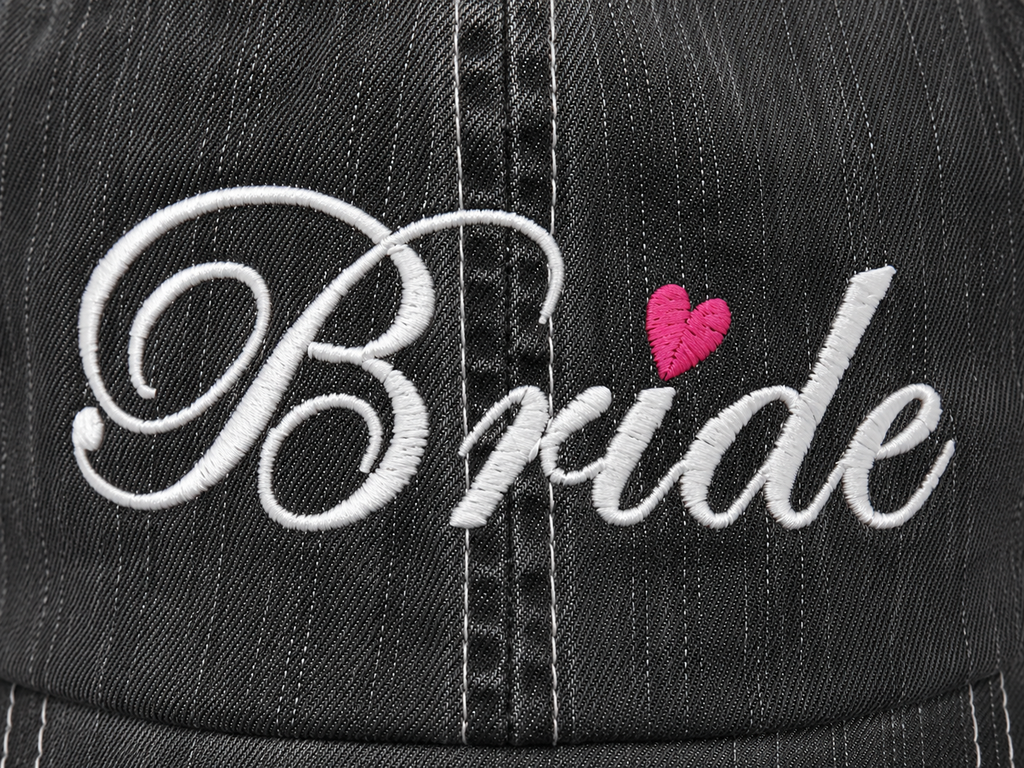 Close-up of white Bride script embroidery with pink heart accent on distressed charcoal trucker hat
