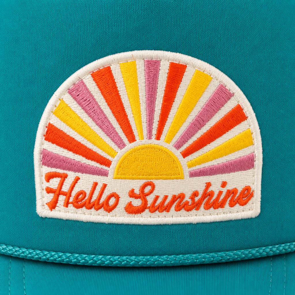 A close-up of a teal cap featuring a colorful embroidered patch with a semi-circular sunburst and the words 'Hello Sunshine' in orange script.