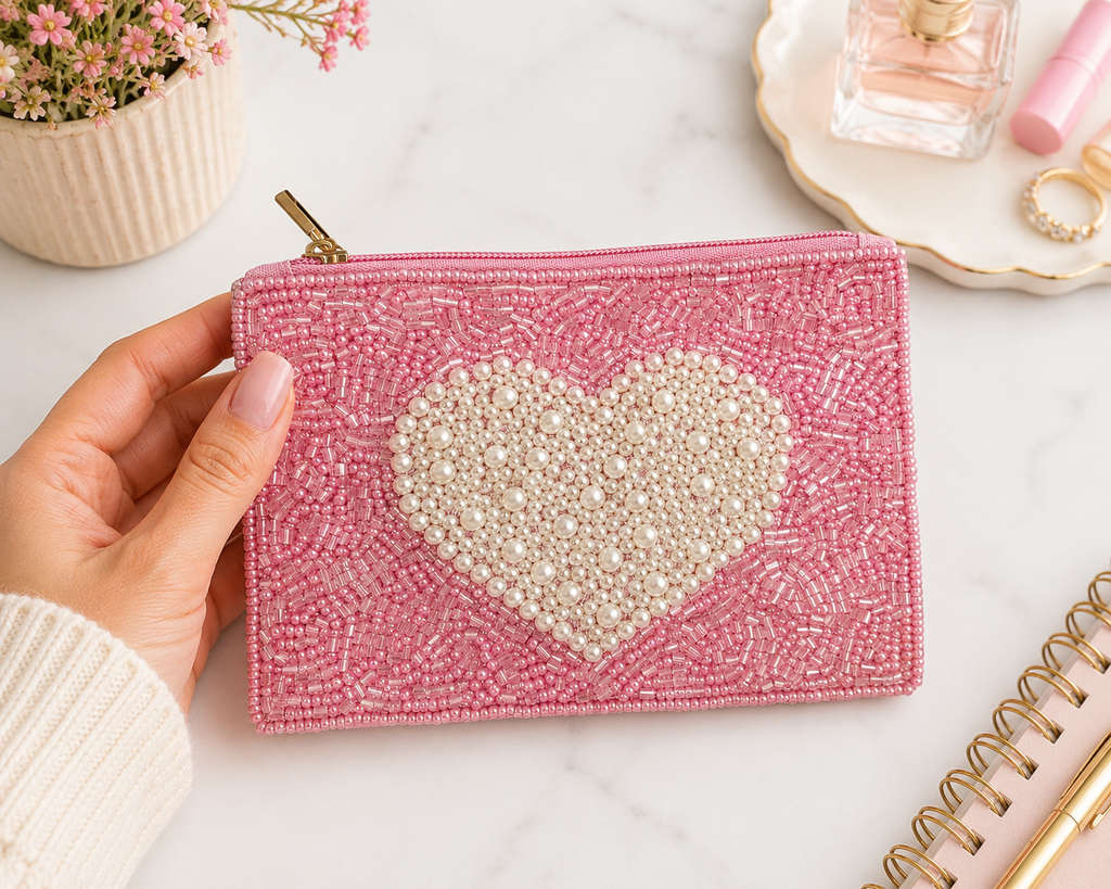 A pink beaded zip pouch with a large white beaded heart on the front, held by a hand over a marble surface with cosmetic props in the background.