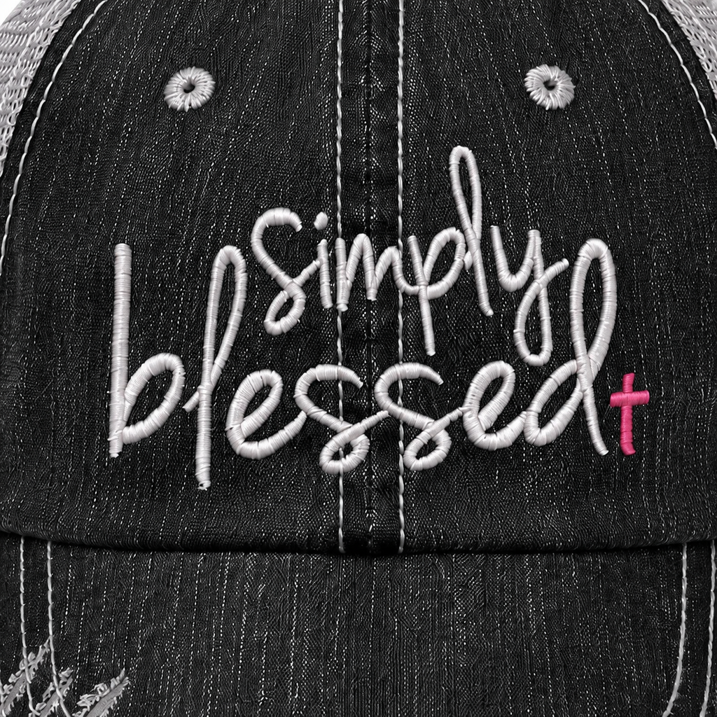 Close-up of a black denim-textured baseball cap with white embroidered script reading 'simply blessed' and a small pink cross accent on the right side.