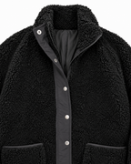 Close-up of the black sherpa jacket showing its stand-up collar, zipper, silver-tone snaps, smooth black placket, and pocket trim
