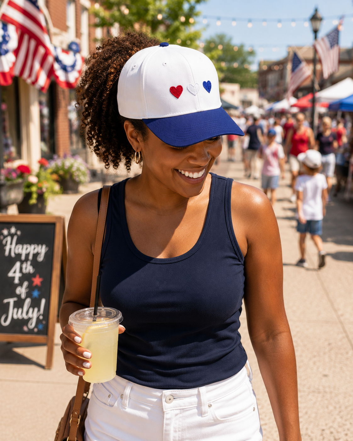 A smiling woman walks through a 4th of July street festival wearing a white and navy Red White & Blue Hearts Baseball Cap, holding a lemonade, with American flags and a Happy 4th of July sign in the background.
