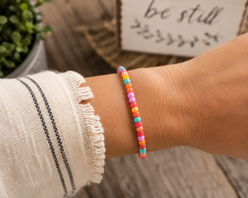 Woman wearing colorful seed bead stretch bracelet on wrist with white fringe top and rustic be still sign backdrop

