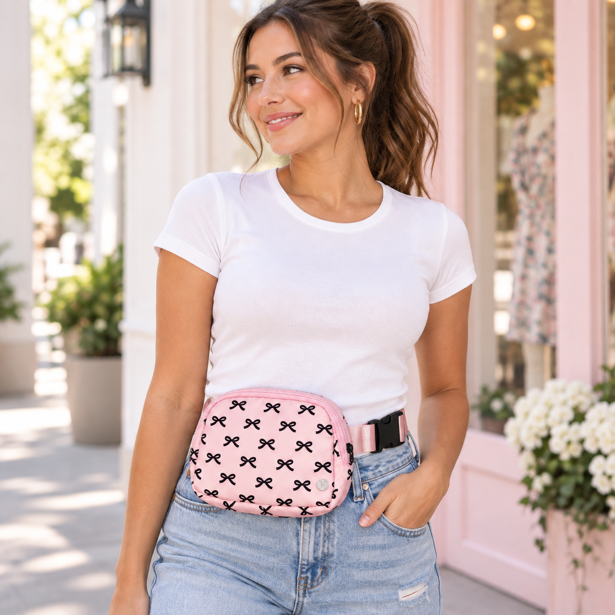 Woman in white tee and jeans wearing Pink Bow Belt Bag at the waist showing pink nylon with all-over black bow print and adjustable pink strap with black buckle in front of bright boutique exterior
