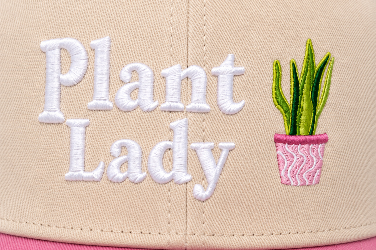 Close-up of white embroidered "Plant Lady" lettering with a detailed potted snake plant embroidery on a cream baseball cap front panel.

