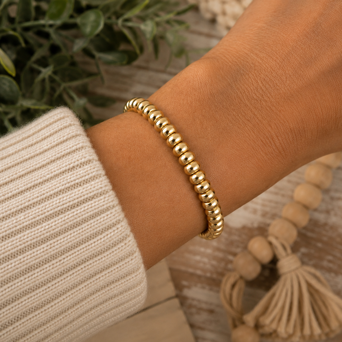 Woman wearing gold beaded stretch bracelet on wrist with cream knit sweater and greenery background
