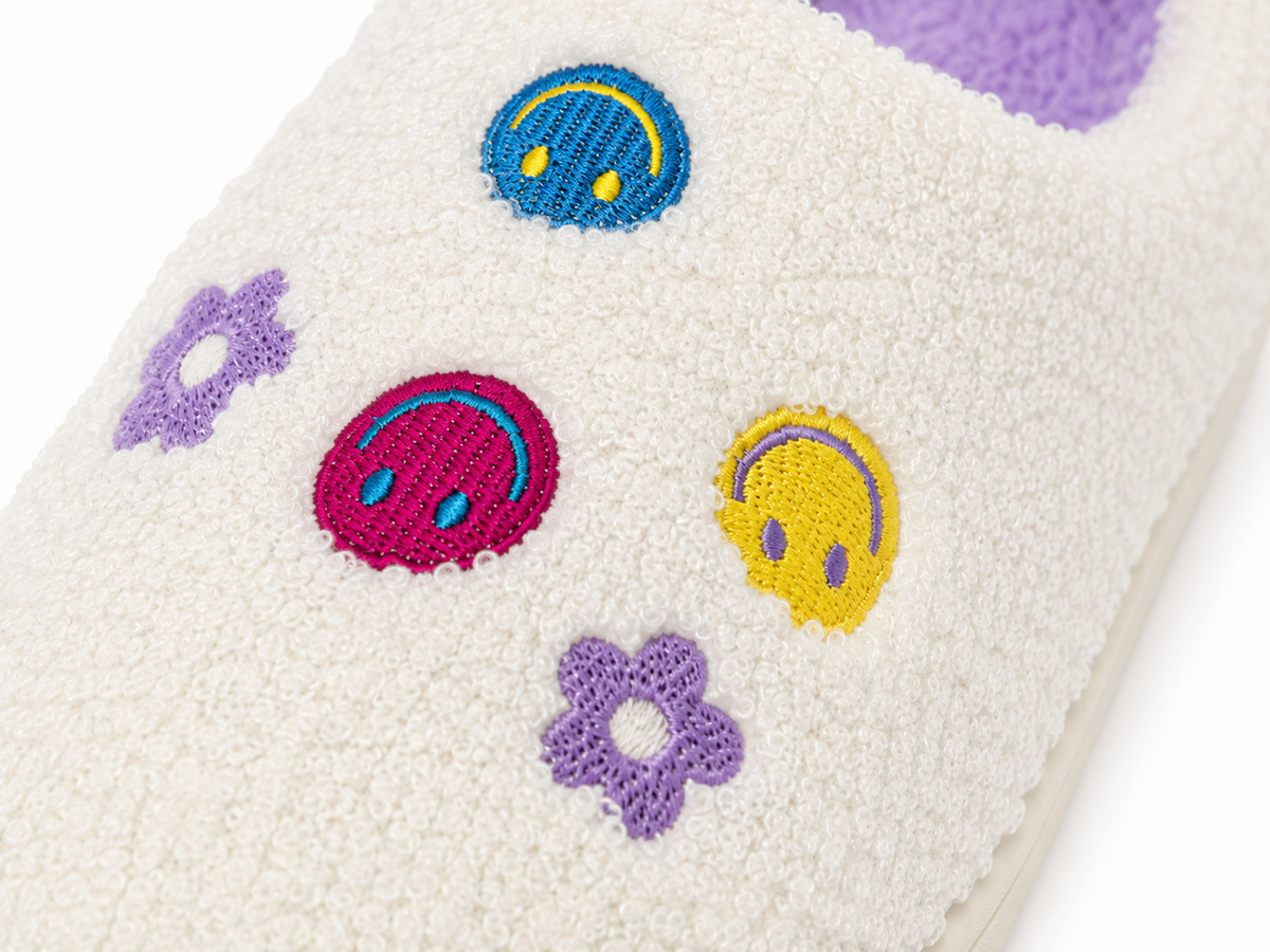 Close-up of colorful embroidered smiley face accents and purple daisy flowers on Ivory Smiley Face Fuzzy Slippers showing blue, pink, and yellow melty smiley face designs on soft ivory fabric
