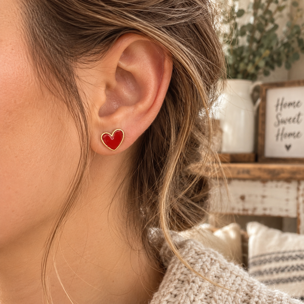 Woman wearing red heart stud earring with gold-tone trim on ear with cream knit sweater and Home Sweet Home sign backdrop
