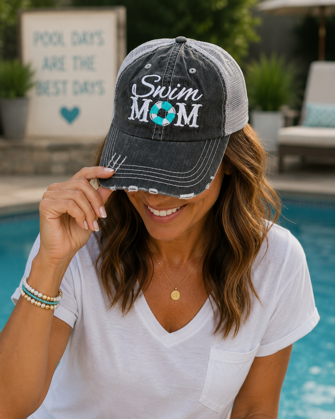 A woman wearing a distressed trucker-style baseball cap with 'Swim MOM' embroidered on the front, posing by a swimming pool.