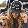 Woman wearing Dog Mother Wine Lover Trucker Hat with distressed black front and gray mesh back showing white embroidered Dog Mother Wine Lover lettering with gray pullover and golden retriever backdrop
