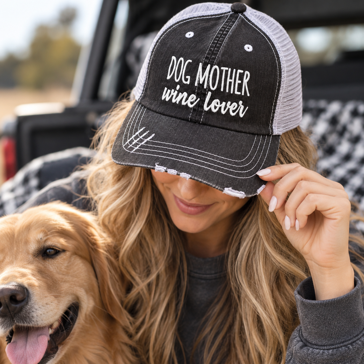 Woman wearing Dog Mother Wine Lover Trucker Hat with distressed black front and gray mesh back showing white embroidered Dog Mother Wine Lover lettering with gray pullover and golden retriever backdrop
