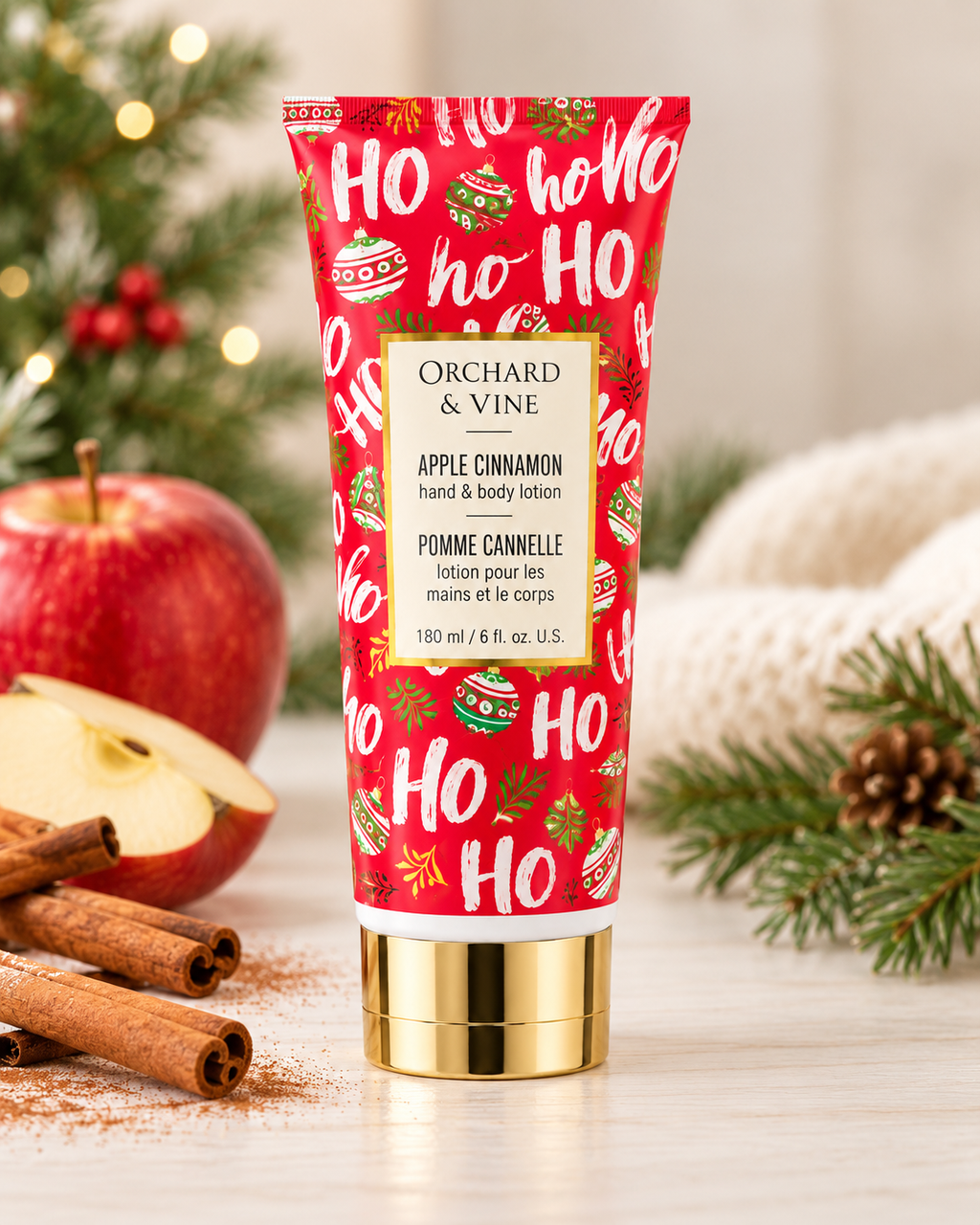 Apple Cinnamon hand and body lotion in a red holiday tube, displayed beside apples, cinnamon sticks, a towel, and Christmas greenery
