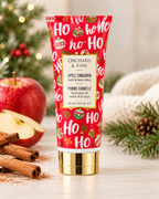 Apple Cinnamon hand and body lotion in a red holiday tube, displayed beside apples, cinnamon sticks, a towel, and Christmas greenery
