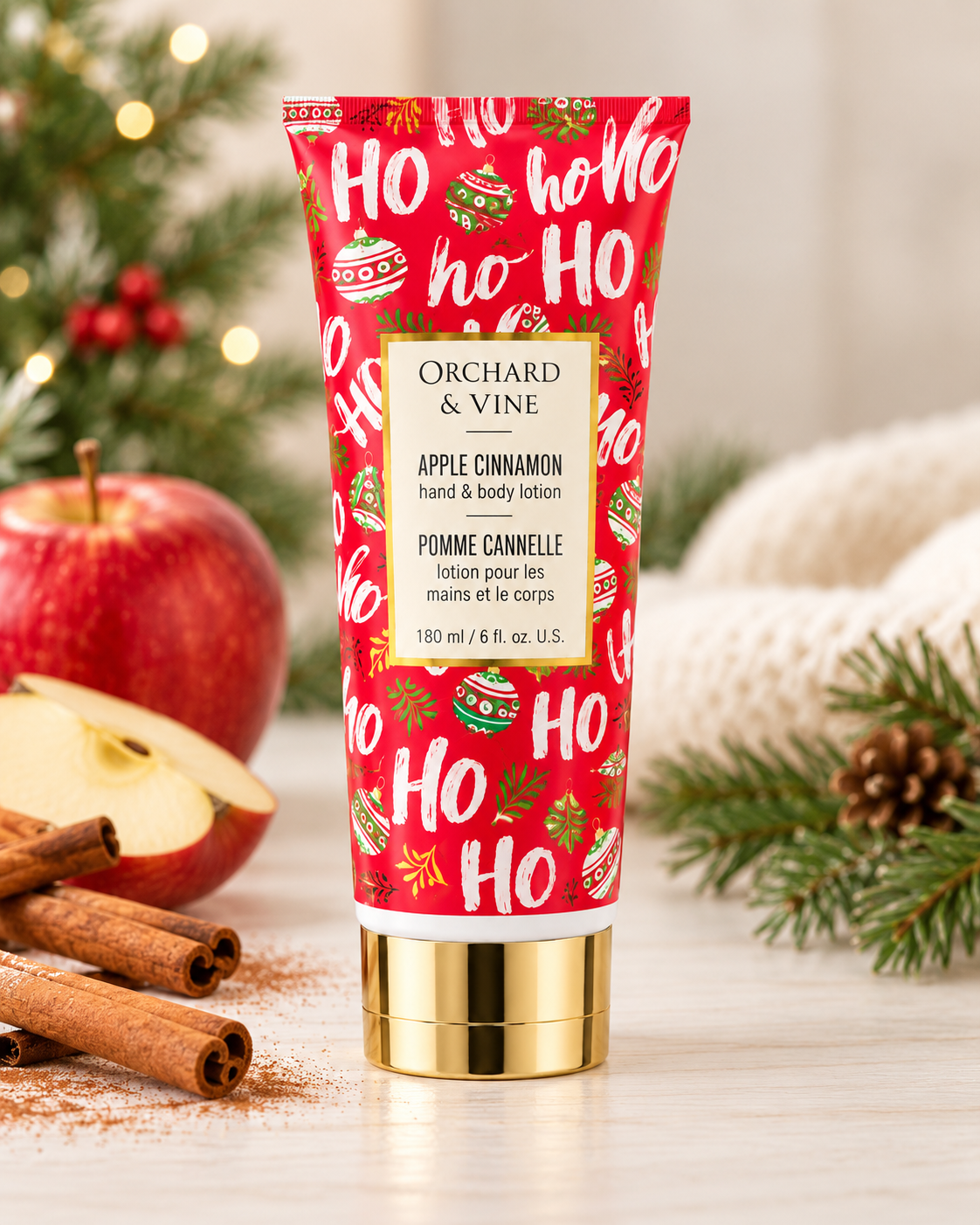 Apple Cinnamon hand and body lotion in a red holiday tube, displayed beside apples, cinnamon sticks, a towel, and Christmas greenery
