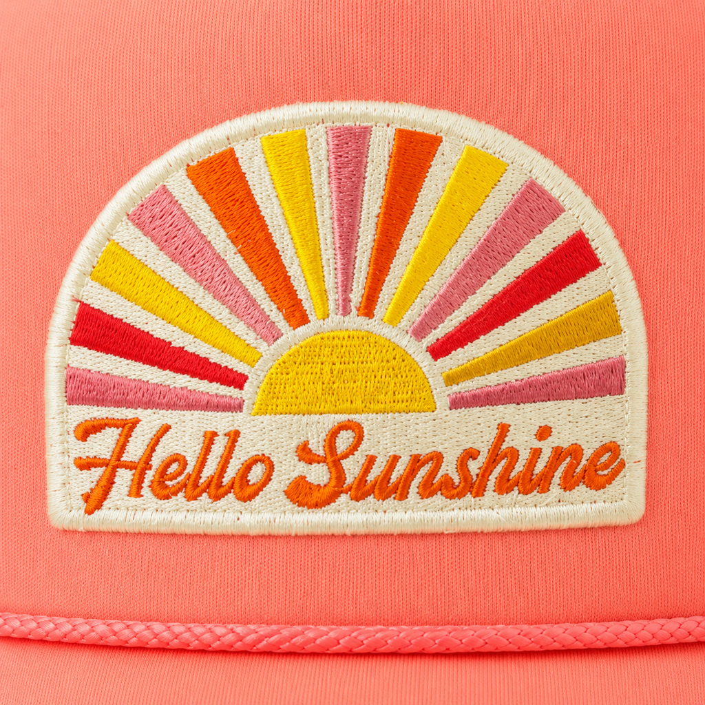 A close-up of a coral pink cap featuring a rectangular embroidered patch with a retro sunburst design in yellow, orange, red, and pink, with the words 'Hello Sunshine' stitched in orange below.