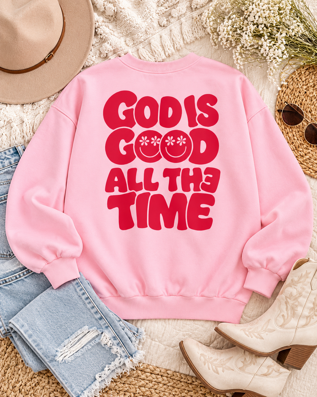 Back of the pink God Is Good sweatshirt styled with jeans, cream cowboy boots, a tan hat, sunglasses, and white flowers
