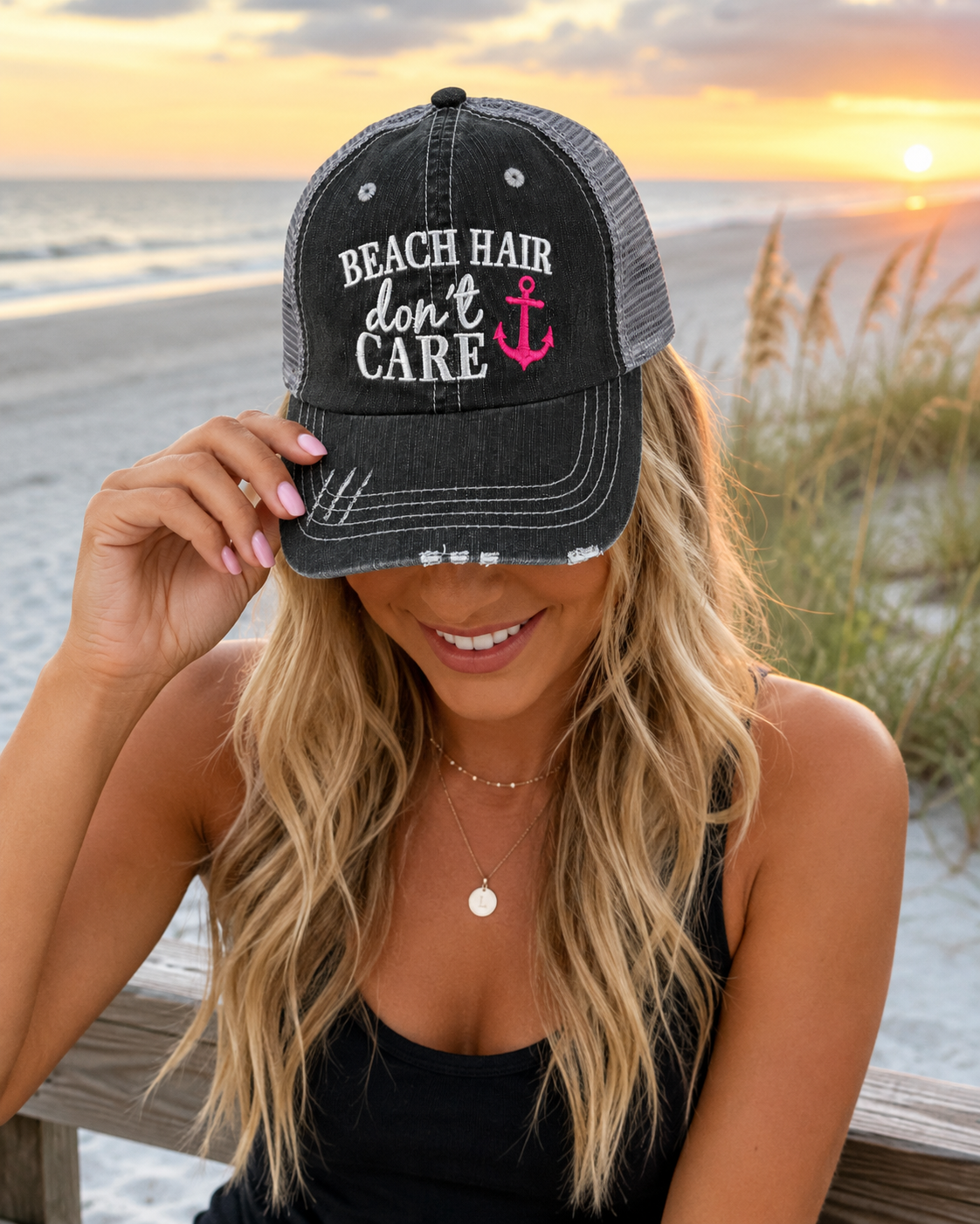 A woman at the beach wearing a distressed black trucker hat with 'BEACH HAIR don't CARE' and a pink anchor graphic on the front. The background shows a sunset over the ocean with sand dunes and beach grass.