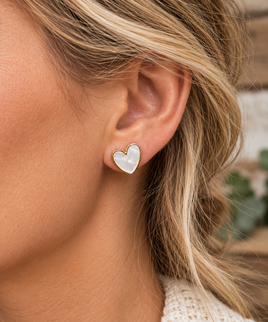 Woman wearing pearl heart stud earring with gold-tone trim on ear with blonde hair, cream knit sweater, and greenery backdrop
