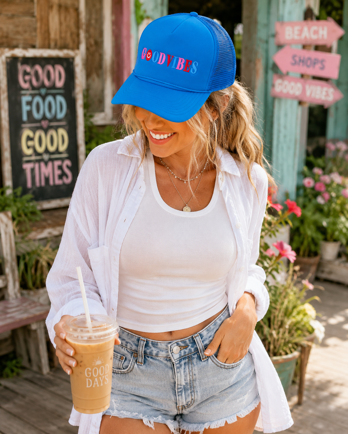 A smiling woman outside a colorful beach shop holds an iced coffee wearing a bright blue Good Vibes Trucker Hat with multicolor embroidered lettering and smiley face accent.
