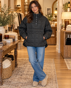 Brunette woman smiling in black quilted puffer pullover jacket with gunmetal snap quarter placket and large kangaroo pouch pocket, paired with wide-leg blue jeans and cream platform slippers in a warm boutique interior with olive tree and wood tables
