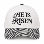 He Is Risen Baseball Cap in Zebra front view showing white cotton crown with black decorative script He Is Risen embroidery, black and white zebra print bill, and zebra print top button on white background

