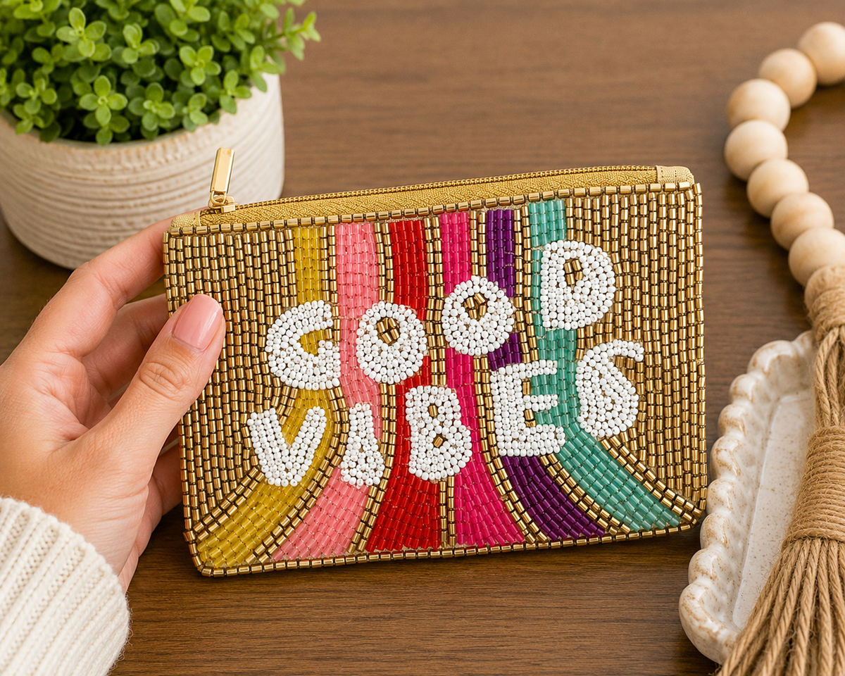 A hand holds a gold beaded zip pouch with colorful vertical stripes and white beaded text reading 'GOOD VIBES' on a wooden tabletop, with a potted plant and decorative beads in the background.