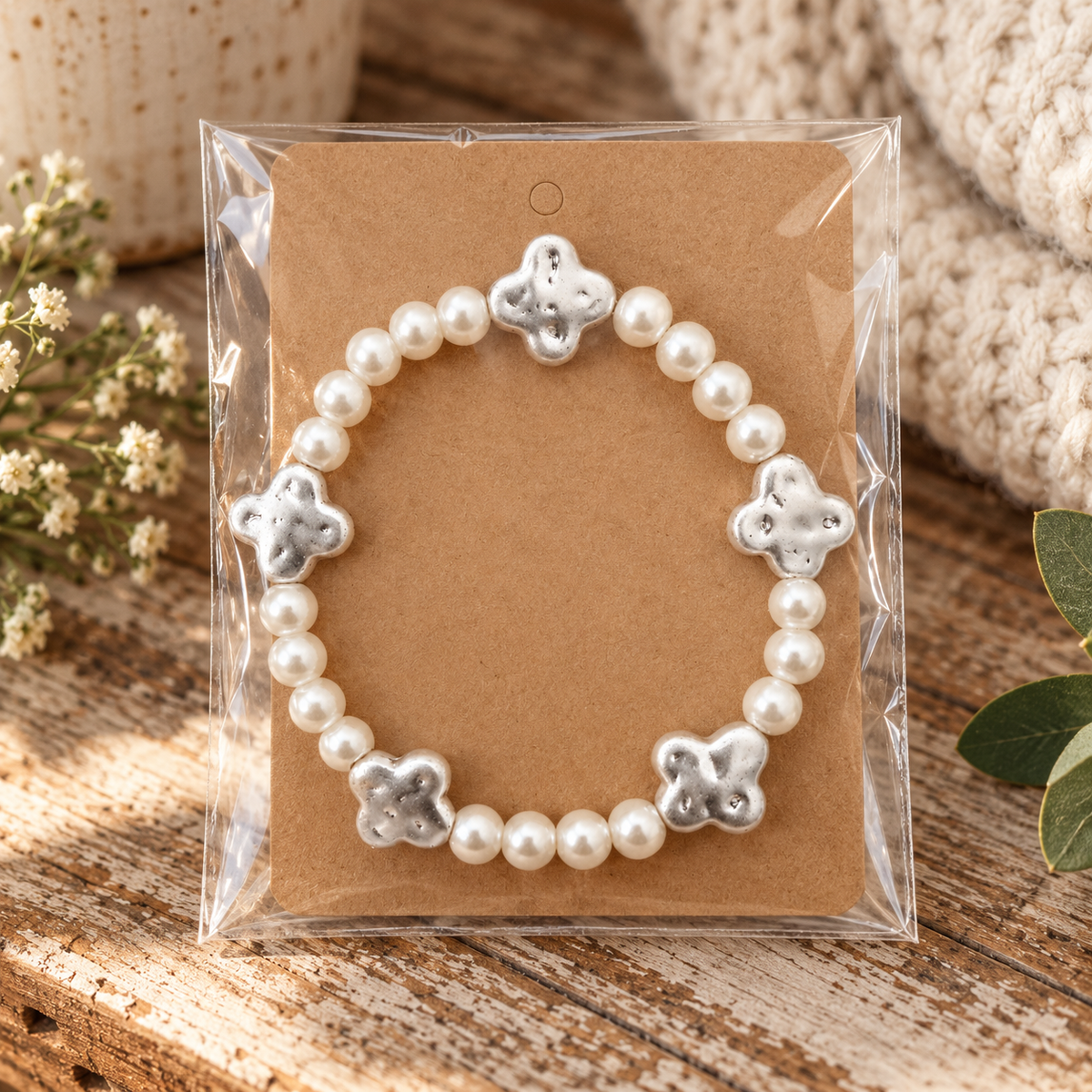 Pearl beaded bracelet with silver clover accents on kraft card in clear packaging on rustic wood surface with white floral sprigs, cream knit, and eucalyptus greenery
