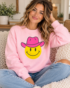 Model wearing the light pink cowgirl smiley sweatshirt with a yellow happy face and hot pink cowboy hat, paired with jeans
