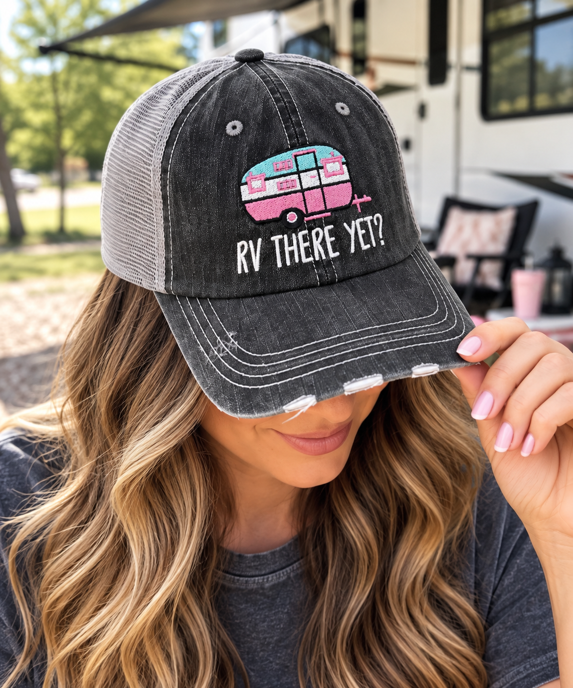 Woman wearing RV There Yet Trucker Hat with distressed black front and gray mesh back tilted down showing aqua and pink embroidered camper graphic with white RV There Yet lettering and RV backdrop
