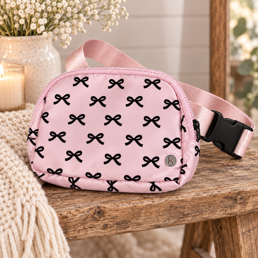 Pink Bow Belt Bag and Crossbody on rustic wood bench showing pink nylon with all-over black bow print, silver Katydid K logo rivet, and adjustable pink satin strap with black buckle beside cream stoneware vase of white flowers and lit glass candle with chunky knit throw on white shiplap background
