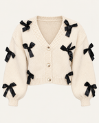 Front view of the tan cropped cardigan showing black ribbon bows, a ribbed V-neckline, three front buttons, and long sleeves
