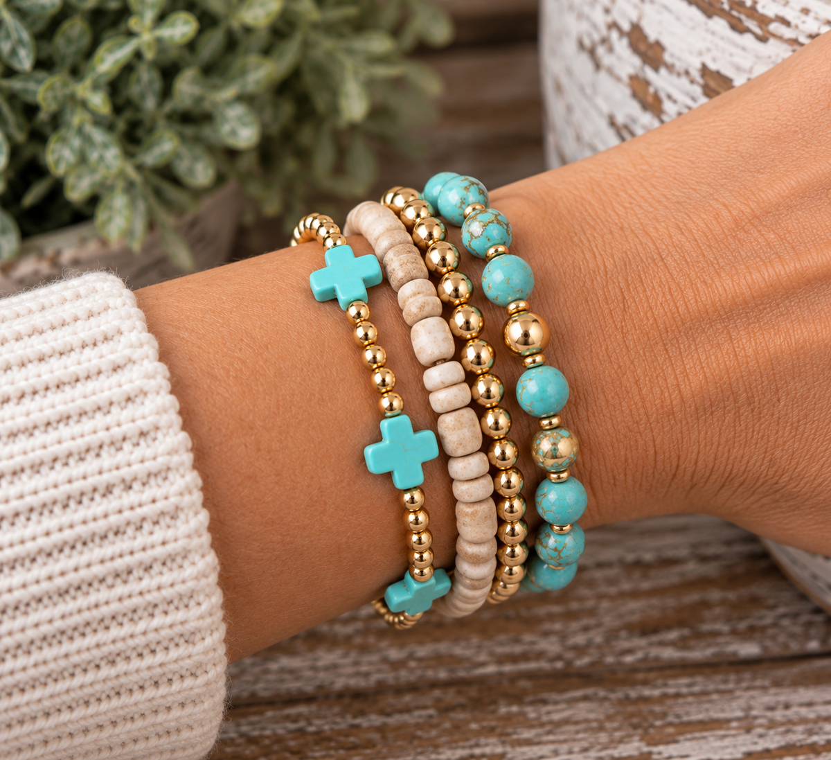 Gold and turquoise cross bracelet stacked with white heishi gold bead bracelet and turquoise stone gold bead bracelet on wrist with cream knit sweater and rustic wood backdrop
