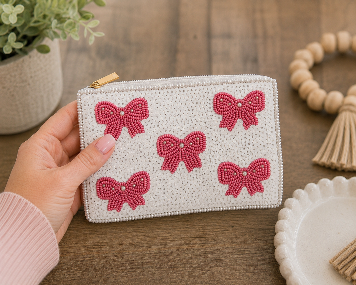 A hand holding a white woven zip pouch decorated with five pink beaded bow motifs, set against a rustic wooden tabletop with decorative props.
