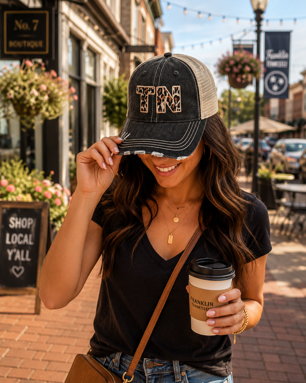 A woman wearing a distressed trucker hat with 'TN' in leopard print on the front, standing on a brick sidewalk in front of a boutique.