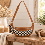 Duo Checkered Crossbody Bag in Camel on rustic wood bench showing black and cream checkered body with camel tan trim and gold hardware beside white ceramic pitcher with dried white florals, lit pillar candle in wicker holder, stacked books, and wood bead garland in wooden bowl on white shiplap background
