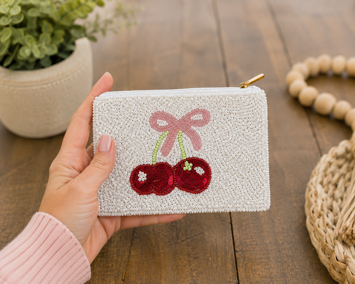 A hand holding a small white beaded zip pouch decorated with a pink bow and red cherry design, set against a rustic wooden table background with a potted plant and woven basket.