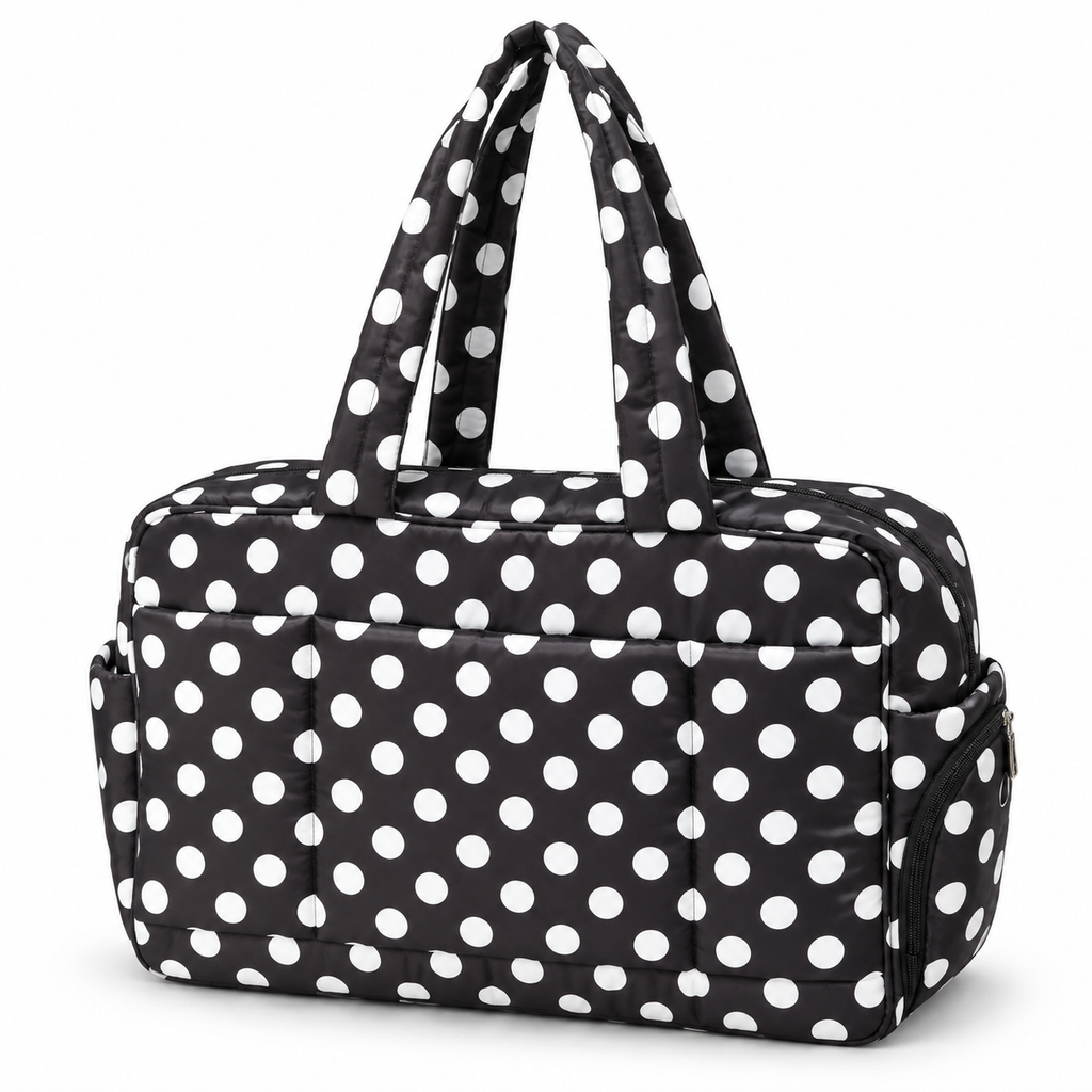 Black Polka Dot Shoulder Tote Bag three-quarter angle view showing black padded quilted fabric with bold white polka dots, dual dot-print shoulder handles, top zipper closure, front slip pocket, and side zippered pocket with silver hardware on white background
