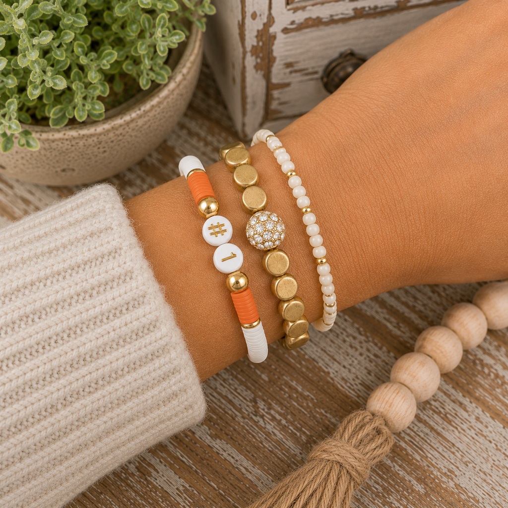 Gold crystal accent stretch bracelet stacked with orange and white #1 bracelet and cream seed bead bracelet on wrist
