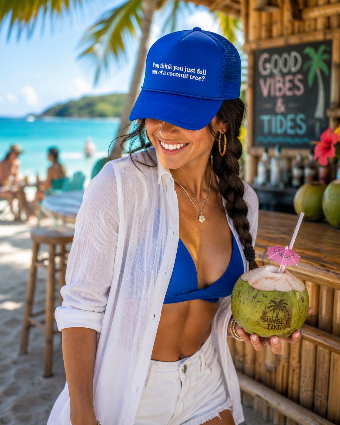 A woman at a beach bar wearing a royal blue Coconut Tree Trucker Hat with white embroidered "You think you just fell out of a coconut tree?" quote, holding a coconut drink.
