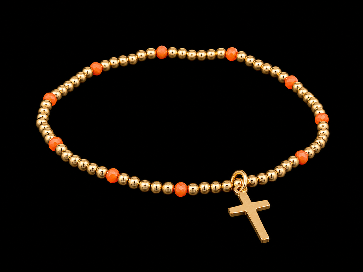Gold beaded stretch bracelet with orange faceted accents and dangling gold cross charm shown at an angle on black reflective surface
