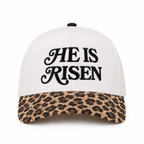 He Is Risen Baseball Cap in Leopard front view showing cream cotton crown with black decorative script He Is Risen embroidery, tan and brown leopard print bill, and black top button on white background

