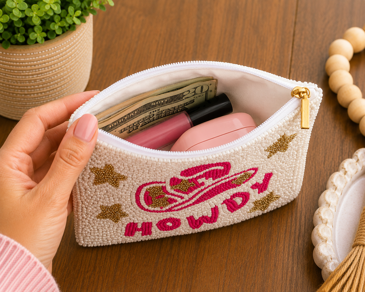 A hand holds an open cream-colored beaded cosmetic pouch with pink and gold embroidery reading 'HOWDY' and a cowboy hat graphic. The pouch is shown on a wooden table with makeup items inside, including a pink lip product and a black pen.