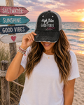A woman at a beach during sunset wearing a distressed black and gray trucker hat with the text 'High Tides Good Vibes' in white, paired with a white t-shirt. A wooden signpost with directional signs reading 'SALTWATER', 'SUNSHINE', and 'GOOD VIBES' stands next to her.