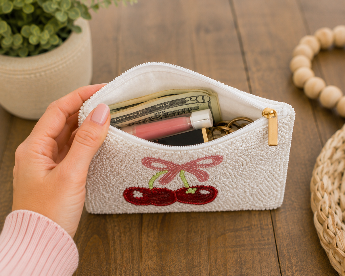 A hand holds an open white textured zip pouch with a cherry embroidery on the front. The pouch contains cash, lip balm, and keys, and is set on a wooden tabletop with a potted plant and woven decor in the background.