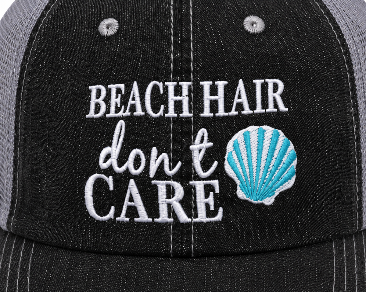 Close-up of white embroidered "Beach Hair Don't Care" lettering with teal seashell accent on a distressed black trucker hat front panel.

