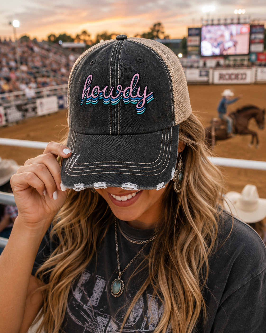 A woman at a rodeo wearing a distressed black trucker hat with tan mesh back and "Howdy" embroidered on the front, with a cowboy on horseback in the background
