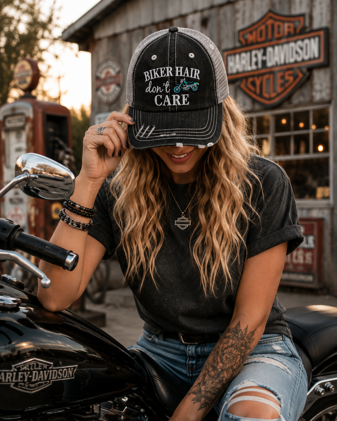 A woman wearing a black and gray distressed trucker hat with the text 'BIKER HAIR don't CARE' sits on a black Harley-Davidson motorcycle in front of a rustic Harley-Davidson shop.